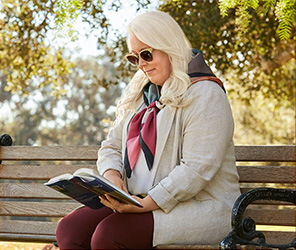 TEPEZZA patient Jeanne T. sitting on a bench reading while wearing sunglasses